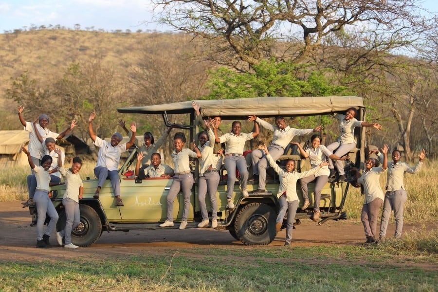 The women of Dunia Safari Camp together around a jeep with their arms raised and happy faces, in Tanzania Africa