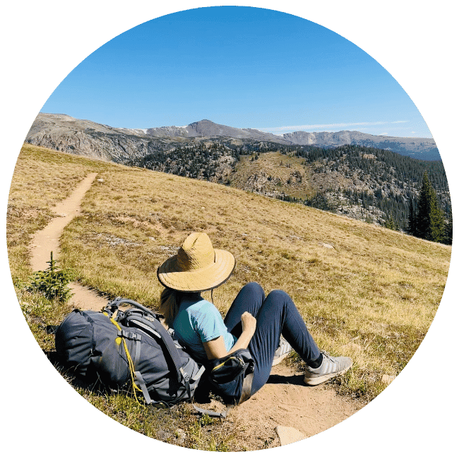 A woman sits on a trail and looks off into the distance toward mountains